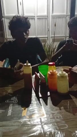 Two young men are seated at an outdoor table, both looking at their phones. A series of condiment bottles, including ketchup, mustard, and mayonnaise, sit in the center of the table. The table is covered with a plastic tablecloth, and some printed materials are visible underneath. A green plastic cup is also on the table. In the background, a white garage door is partially open, with a white dog lying near the doorway.