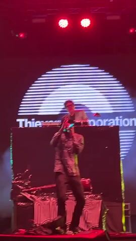A male singer wearing a patterned shirt and a hat stands center stage, holding a microphone and performing. To his right, another male with dreadlocks is dancing. Behind them, a man in a suit stands at a DJ booth with turntables, and a large screen displays a logo with concentric circles and text. The stage is lit with red and green lights, and smoke effects are visible. The overall environment appears to be a live music performance.