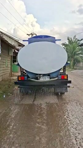 From a low-angle, the rear of a blue-and-white water truck is shown, driving down a dirt road. The truck has a large, oval-shaped water tank and a black bumper with a system of pipes that are spraying water onto the road. The water spreads across the road's surface, creating a wet and muddy appearance. On either side of the road, there are simple, one-story buildings with wooden exteriors. The building on the left has a green door. A tall palm tree is visible to the right. Overhead, power lines stretch across the sky, which is filled with large, puffy white clouds. The sky is a mix of blue and grey, suggesting a partly cloudy day. The truck's license plate is visible, reading "BL 8246 NW." The camera remains static as the truck moves away, continuously spraying water.