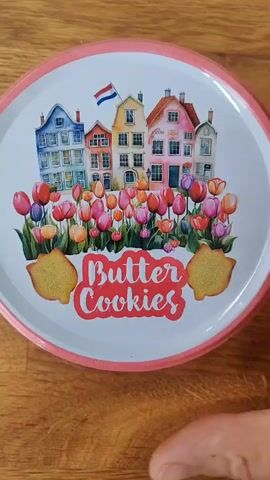 A circular tin of butter cookies is opened on a wooden surface. The tin has a colorful lid depicting Dutch houses and tulips, along with the words "Butter Cookies". Inside, the cookies are arranged in a decorative manner, separated by fluted paper liners. The video shows the lid being removed, revealing the cookies, and then close-ups of the cookies themselves.