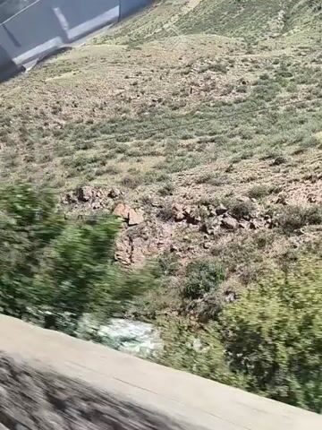 The video shows a high-angle view of a rocky, scrub-covered hillside with a body of water at its base. The water appears to be flowing, possibly a river or stream, and is visible on the left side of the frame. The hillside is characterized by sparse vegetation, primarily green and brown shrubs and grasses, interspersed with rocky outcrops and patches of bare earth. The terrain is uneven and slopes upwards away from the water. The lighting suggests it is daytime, with sunlight illuminating the landscape. The perspective suggests the viewer might be in a vehicle moving alongside the hillside, as indicated by a blurred concrete barrier or road edge in the foreground. The overall impression is of a natural, somewhat arid landscape.