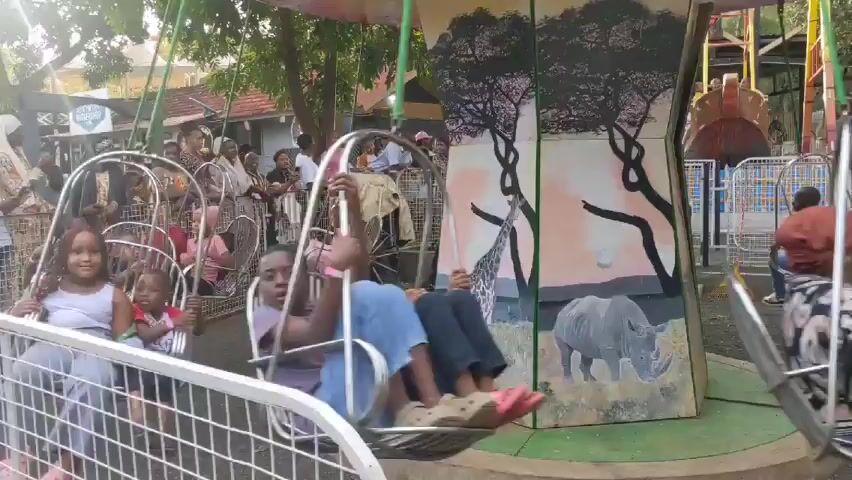 The video shows a group of children and adults riding on a swing ride at an outdoor amusement park or fair. The swings are suspended from a rotating platform, and the riders are seated in individual chair-like swings. A crowd of onlookers is visible behind a metal fence, watching the ride. The backdrop of the ride features a painted African savanna scene with a giraffe and a rhinoceros. The overall atmosphere seems lively and festive.