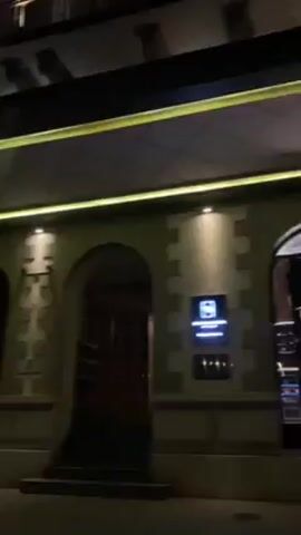 The video shows a building at night. The building has a stone facade with arches. There are lights illuminating the building, including string lights under the awning and lights on the windows. The building has multiple stories, and the windows are lit up. There is signage on the building.