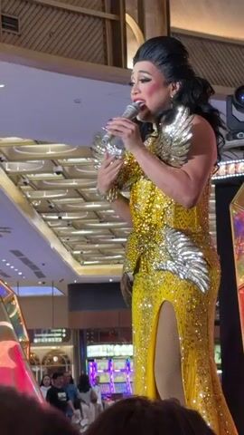 A drag queen in a yellow sequined gown with gold wing-like adornments on the shoulders and waist performs on stage, holding a microphone and gesturing with her left hand. The background shows a brightly lit interior with a sloped ceiling, what appear to be slot machines, and some reflective decorative elements. The lighting is theatrical, with green spotlights visible.