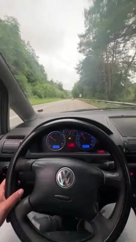 The video provides a first-person perspective from the driver's seat of a Volkswagen vehicle, likely in Schopp, Germany. The driver's hands are visible on the black steering wheel, which features the silver VW logo in the center.

Through the windshield, an asphalt road stretches ahead, flanked by dense green trees on both sides. The sky is overcast, suggesting cloudy weather. Several other cars are visible in the distance on the road.

The car's dashboard is prominent, with the speedometer and tachometer featuring blue backlighting. A digital display between these gauges shows "10:12" for the time and "16.0°C" for the temperature. Several red and orange warning lights are illuminated on the dashboard, including what appears to be a battery light, an oil light, and a check engine light. The fuel gauge and temperature gauge are also visible. The rearview mirror is positioned at the top center of the windshield. The overall scene depicts a steady drive on a road, likely a main road or highway, surrounded by natural scenery.