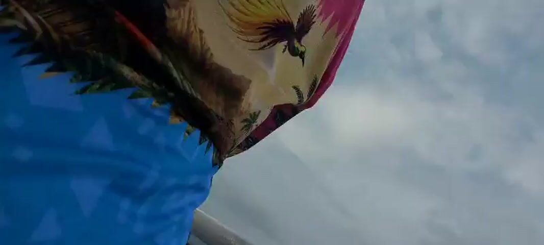 A person is lying on their side, covered with a patterned blanket, in what appears to be a boat. The blanket has a large, colorful image of a dragon. The person is wearing a light-colored hoodie. The camera is positioned above and to the right of the person, looking down at them. The scene is brightly lit, and the background is a mix of blurry colors. A large, white boat cabin with windows is visible in the distance. The sky is cloudy.