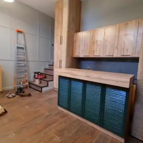 A wide shot captures a partially finished kitchen area and a staircase. To the right, a kitchen island with a wooden countertop and green louvered cabinet doors is visible. Above the island are wooden cabinets with dark handles. To the left, a staircase with dark treads and risers leads upwards. Tools, including a ladder and a power drill, are present on the staircase. A tall, narrow cabinet with a wood-grain finish stands between the kitchen and the staircase. A rough-textured, cylindrical object, possibly a cat scratching post, is positioned next to the tall cabinet. The floor is covered with wooden planks. The walls are painted in a neutral gray tone, with some areas featuring decorative paneling.