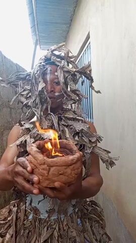 A person is holding a small fire in their hands. They have white paint on their face and are wearing what appears to be a costume made of dried leaves. The person seems to be blowing on the fire. They are standing in an alleyway or narrow outdoor space with a concrete wall on one side and a building with a window on the other. The sky is visible above.