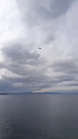The video shows a wide body of water under a cloudy sky. The water is a dark, muted blue-gray and appears calm with small ripples. In the distance, a dark, low-lying landmass with hills or mountains is visible along the horizon. The sky is filled with heavy, gray clouds, suggesting overcast weather or impending rain. A small, dark object, possibly a bird, is seen flying in the upper left quadrant of the frame. The foreground shows a small section of a pebble beach. The video is shot from a stationary perspective, looking out over the water.