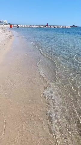 A bright, sunny day at a beach. The camera pans across the shallow, clear water as gentle waves lap the sandy shore. In the distance, a breakwater extends into the sea, with two small lighthouses visible at its end. The water is so clear that the sandy bottom is visible beneath the surface, with sunlight creating shimmering patterns of light and shadow. The sky is a clear blue, with no clouds in sight.