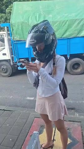 A person wearing a helmet and a pink skirt is standing on a sidewalk looking at their phone. A large truck with a green tarp is driving down the road. A person on a motorcycle is also driving down the road. The scene takes place during the day, and the weather appears to be warm. The person on the sidewalk has long hair braided into a ponytail. They are wearing a white jacket over a light-colored shirt and a light-colored skirt. They are also wearing flip-flops. The truck is blue and has a green tarp covering its cargo. The motorcycle is black. The road is paved, and there is a sidewalk on the right side of the road. There are trees and buildings in the background.