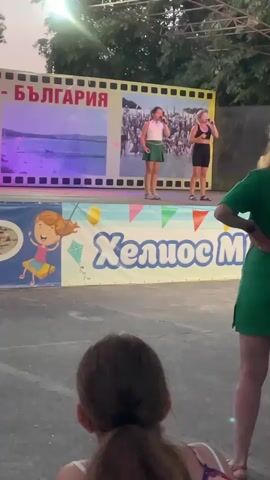 The video shows a performance on an outdoor stage. Two young girls are singing into microphones on the stage. One girl is wearing a white shirt and a green skirt. The other girl is wearing a black tank top and black shorts. Behind them is a large banner with images and text that appears to say "BULGARIA" in large red letters, along with other images and text in Bulgarian. Below the stage, there is another banner with a cartoon of a girl and text that says "Хелиос Ми" (Helios Mi). In the foreground, the back of a young girl's head and shoulders are visible, and to her right, the back of a woman wearing a green shirt is also visible. The background shows trees and what appears to be stage lighting equipment. The overall scene suggests a public event or festival.