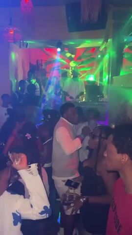 The video shows a crowded indoor party scene with vibrant, colorful lights, primarily blue, red, and green, illuminating the space. Many people are visible, dancing and socializing. A person at the back operates a DJ setup. In the foreground, people dance and interact. One person holds a hookah pipe. Drinks and cans are on a yellow barrel on the floor. The overall atmosphere is energetic and lively.