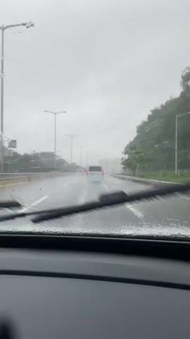 This video shows a view from inside a car driving on a wet road during a rainstorm. The windshield wipers are intermittently clearing the view of the road ahead, which is lined with streetlights and trees on the right side. Several other vehicles are visible on the road, including a white van in the distance. The weather appears overcast and gray.