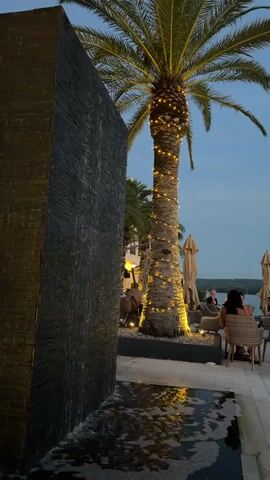 The video shows an outdoor dining area at dusk, with a fountain on the left and a palm tree wrapped in lights in the center. Several tables and chairs are arranged on a tiled patio, with umbrellas providing shade. People are seated at some of the tables, appearing to be dining or socializing. The sky is a deep blue, indicating the evening hour. The overall ambiance is relaxed and possibly luxurious, given the lighting and the waterfront setting.