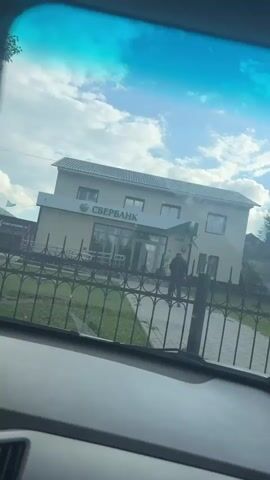 A shot from inside a vehicle looking out at a building with a sign that reads "SBERBANK" in large green letters. The building is light-colored with a dark roof. There is a fence in front of the building, and a person is standing near the entrance. The sky is blue with some clouds.