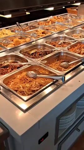 The media file shows a close-up, horizontal panning shot of a buffet food display. The scene is set in a restaurant or eatery, likely a self-service establishment given the array of food trays. The lighting is bright and artificial, suggesting an indoor setting, possibly during the day or evening.

The buffet features numerous stainless steel trays filled with various dishes. These include what appears to be fried rice, noodles, stir-fried vegetables, various meats in sauce, and seafood, such as shrimp and mussels. Serving utensils are present in many of the trays, indicating that customers are meant to serve themselves. Below the food display, stacks of white plates are visible, ready for use.

The setting appears to be clean and well-maintained. The focus is entirely on the food, with no people visible. The time of day and weather are not discernible from the image. The location context of "Salt, Spain" does not provide any direct visual cues within the media file itself, but it might suggest the type of cuisine or clientele. No visible text is present in the media file.