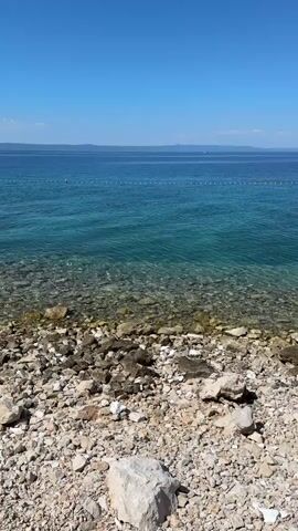 This is a vertical, stable shot of a rocky beach with clear turquoise water. The water is shallow near the shore, revealing the rocky seabed. In the distance, a line of buoys marks a boundary in the water, and beyond that, the horizon is visible with a hazy landmass. The sky is a clear, bright blue with a few wispy clouds. The foreground is dominated by various sizes of light-colored rocks and pebbles. There are no people or animals visible in the frame. The overall impression is one of a peaceful, natural coastal environment on a sunny day.
