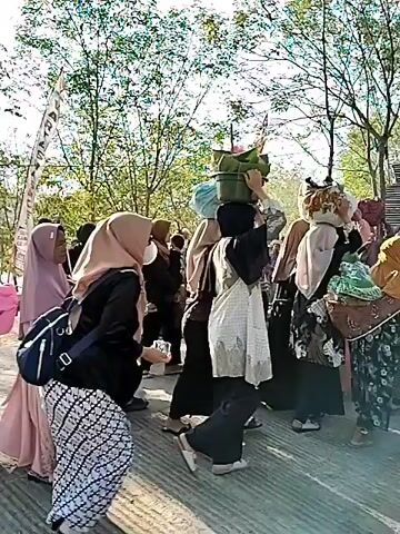 A group of people, mostly women wearing hijabs and long dresses, are walking along a path lined with trees. Some are carrying bags and baskets on their heads or in their hands. The sun is shining, casting shadows on the ground. One woman in the foreground is carrying a rolled-up mat over her shoulder, and another woman in a pink headscarf is carrying a large pink bag. The overall atmosphere appears to be that of a procession or a gathering.