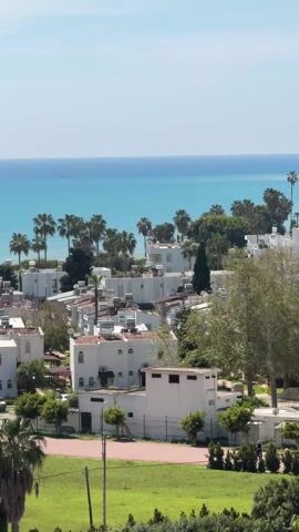 The video shows a daytime exterior view of a cluster of white buildings with palm trees, set against a backdrop of a turquoise sea and a clear blue sky. The buildings are mostly two-story structures with flat roofs, some featuring what appear to be solar panels. Trees and green spaces surround the buildings, and a patch of green lawn is visible in the foreground. The ocean is calm, with a very small boat visible in the distance.