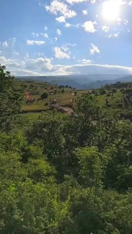 A drone shot pans over a lush green landscape under a bright, sunny sky with scattered clouds. In the distance, a valley with rolling hills and mountains stretches out. In the middle ground, a group of people are gathered on a grassy slope, some standing and some walking. A vehicle, possibly an off-road vehicle, is visible on a dirt path. Red flags are planted along the path, suggesting a race or event. The foreground is dominated by the dense foliage of trees and bushes.