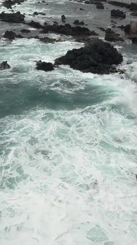 The media file captures a dynamic scene of the ocean crashing against a rocky shoreline in Puerto de la Cruz, Spain. The primary subjects are the churning, turquoise-colored water and the dark, jagged rocks that punctuate the coastline. In the background, a concrete walkway or promenade is visible, with several small figures of people scattered along it, suggesting a public area. Some have railings or structures, indicating a developed viewing point.

The main activity is the powerful surge of waves breaking and foaming against the rocks, creating a dramatic display of nature's force. The water is a vibrant, almost electric blue-green, contrasting with the stark black of the volcanic rocks. White seafoam churns and spreads across the surface, particularly in the foreground, obscuring some of the submerged rocks. The lighting suggests it is daytime, possibly overcast, as there are no strong shadows, and the light is diffused. The overall atmosphere conveyed is one of raw, untamed coastal beauty and the relentless energy of the sea. The location cues of volcanic rocks and the specific mention of Puerto de la Cruz, Spain, place the scene in a well-known tourist area with a distinctive geological landscape. There is no discernible text visible in the media file.