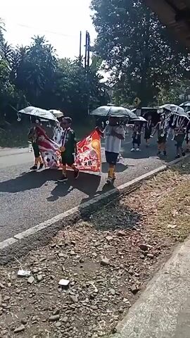 A group of people, some holding umbrellas, are marching along a paved road. The people appear to be young, possibly children or teenagers, and are wearing matching uniforms. Some are carrying a banner that appears to be red and yellow. Several of the umbrellas are decorated to resemble soccer balls. The road is lined with vegetation, including trees and bushes. The sky is clear and sunny, suggesting it is daytime. The overall scene appears to be a parade or procession.
