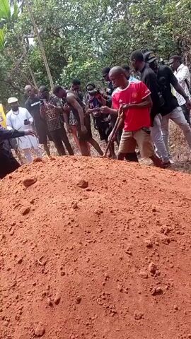 The video shows a group of people gathered around a large mound of red earth, likely at a burial site. A person in a red Manchester United jersey is using a shovel to move earth, apparently helping to fill in a grave. Another person dressed in black is also shoveling dirt into the hole. Several other individuals are standing around, some dressed in casual attire and others in what appears to be religious clothing, including a nun in a white habit with a blue veil. The scene is set outdoors, surrounded by trees and vegetation. The overall tone suggests a somber occasion.