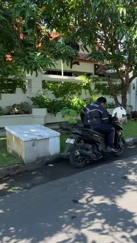 A person wearing a black jacket with white accents and a white helmet is seated on a black motorcycle. The motorcycle is parked on the side of a paved road, adjacent to a grassy area. Behind the grassy area is a building with white walls and a tiled roof, surrounded by trees and lush greenery. A concrete structure, possibly a bench or a planter, is visible in the foreground to the left. The lighting suggests it is daytime, with shadows cast by the trees on the road.