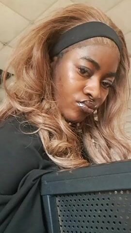 A dark-skinned woman with wavy blonde hair, a black headband, and lip gloss poses for the camera. She holds a dark object with perforations. She looks downward, then directly at the camera, and turns her head slightly. She wears a black top and a delicate gold chain. The background is blurry and appears to be a brightly lit interior.