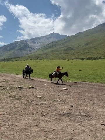 The video shows two people riding horses on a dirt path in a mountainous, grassy landscape. The person on the right is wearing an orange shirt and a hat and appears to be waving. The person on the left is wearing a hat and is further back on the path. The path leads through a vast green field with a mountain range in the background. The sky is blue with scattered clouds. The overall impression is one of an outdoor adventure or journey in a scenic natural setting.