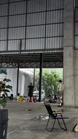 Two men stand in a covered outdoor area, possibly a car wash or garage. The man on the left, wearing a green t-shirt and brown shorts, stands facing away from the camera with his hands on his hips. The man on the right, wearing a light-colored t-shirt and shorts, stands facing a wall with his back to the camera and appears to be looking at something in his hand. In the background, there are several vehicles, including a white car, a yellow scooter, and other motorcycles. A folding chair is positioned in the foreground. The floor is wet, indicating recent rain or washing. The structure has a metal roof and concrete pillars. Lush green foliage is visible beyond the covered area.