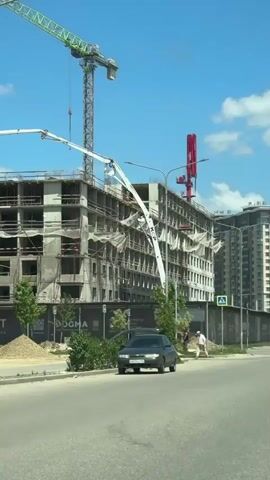 A long vertical shot of a construction site shows an unfinished building with exposed concrete and rebar against a bright blue sky with scattered clouds. A large green and grey tower crane is visible on the left, and a smaller red crane is on the right. A concrete pump with a long white boom extends from the bottom left, over the building, and toward the right side of the screen. In the foreground, a dark-colored sedan is parked on the side of a paved road. Green foliage lines the road, and two blue pedestrian crossing signs are visible. A man in a white shirt and dark pants walks along the sidewalk next to the road. The building has multiple floors, with some sections covered in a light-colored netting. Signs with text are visible at the base of the building, and construction materials are piled on the ground in front of it.