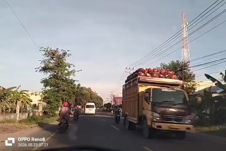 The camera, likely dashboard-mounted, faces forward down a road running through a populated area. On the right of the road is a collection of low buildings, power lines and utility poles. A white and red telecommunication tower is visible, also on the right side of the frame. There are pedestrians standing along the right shoulder of the road. On the left of the road is a collection of trees, bushes, and some buildings. Traffic includes motorbikes and a white van. The sky is light blue and sunny. A timestamp on the lower left corner of the frame indicates 2026.05.23 07:21.