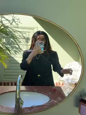 A woman takes a selfie in a round mirror in a bathroom. The woman is wearing a black jacket with gold buttons, and her hair is dark and shoulder-length. She is holding her phone in front of her face. The mirror is mounted on a light green wall. Below the mirror is a sink with a marbled counter. A plant is partially visible in the reflection of the mirror. Light streams in through a doorway or window reflected in the mirror.