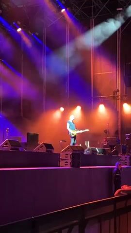 The video shows a live concert performance. A male musician, appearing to be the singer and guitarist, stands center stage playing a bass guitar. He is wearing a dark blue t-shirt and jeans. The stage is equipped with multiple large speakers and lighting rigs. Purple and orange lights illuminate the stage, and smoke or fog machines are in use, creating atmospheric haze. The musician is standing on a raised platform made of stacked speaker cabinets. In the foreground, the edge of a crowd barrier and glimpses of audience members are visible. The camera is positioned in the audience, looking towards the stage.