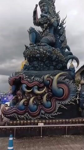 The video shows a large, dark blue statue of a seated figure with elaborate headdress and jewelry. The statue is mounted on a tiered base decorated with numerous skulls and other grotesque figures. The base also features swirling, flame-like elements in blue and red. The statue is outdoors, with a cloudy sky visible in the background. To the left of the statue, there are blurred lights and what appears to be a building or structure. The statue itself is the central focus, with intricate details on its clothing, ornamentation, and facial features. The overall impression is one of a powerful, possibly fearsome, mythical or religious figure.