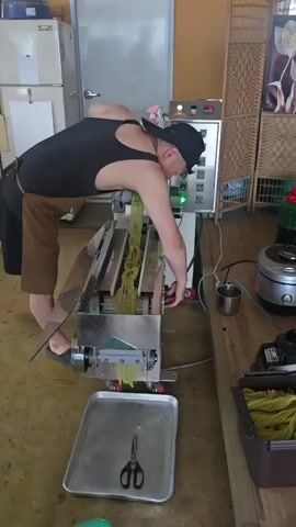 A man wearing a black tank top, brown apron, and black baseball cap is operating a noodle-making machine. He is standing to the left of the machine, bent over as he feeds a yellow noodle mixture into the top. The machine is made of stainless steel and has a control panel with several buttons and lights. Yellow noodles are being extruded from the bottom of the machine and are falling into a metal tray. Scissors are lying in the tray. The scene takes place in what appears to be a commercial kitchen or food production area. There are various appliances and shelves in the background.