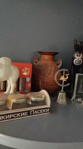 This is a close-up, static shot of a shelf filled with various decorative items. The shelf itself is a light gray, and it's set against a darker gray wall. The lighting suggests an indoor environment, likely during the daytime given the brightness but without any specific clues about the time of day or weather outside.

The collection of objects is diverse and appears to be souvenir-like. On the far left, there's a white, possibly ceramic, camel figurine. To its right is a red box containing what look like small, dark cylindrical objects, possibly incense or candles, and some sparkly material. Next to that is a large, ornate, brown vase with a relief design of figures.

Further to the right, a small silver bell sits on a stand, topped with a golden camel and a crescent moon symbol. A black statue of a cat, reminiscent of ancient Egyptian iconography, stands tall, adorned with a colorful, jeweled collar. In front of the cat is a clear, acrylic replica of the Eiffel Tower.

To the right of the cat statue is a decorative plate featuring a detailed embossed image of a building, labeled "PETRA JORDAN". Behind this, a pyramid-shaped box with intricate carvings is visible. Above the pyramid, a decorative basket with a black and white pattern is topped with a brown, knitted cap or fabric.

Some text is visible on the lower left of the frame, on a white and brown box. The visible portion reads "ОРСКИЕ ПАСЕКИ". The overall impression is of a personal collection, possibly souvenirs from various travels, displayed on a shelf in a home. There are no people or animals in the frame, and the scene is static and still.
