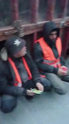 The video shows several construction workers taking a break. They are squatting and sitting along a concrete surface next to some red metal construction forms. Some are wearing high visibility vests and appear to be smoking or using cell phones. Some have material pulled over their heads. The camera moves across the scene, filming the group. The weather appears overcast or cold.