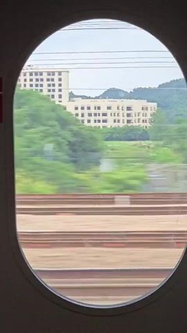 The video captures a view from an oval-shaped train window as it travels through a landscape in Wuxing District, China. The scene transitions between stationary buildings, lush green trees, and passing trains.

Initially, a low-rise building with multiple floors and balconies is visible in the background, situated behind a single-story structure with a row of windows. Train tracks are prominent in the foreground, suggesting movement.

As the video progresses, a tall, modern skyscraper appears, contrasting with the surrounding greenery and older architecture. The speed of the train is evident as trees and buildings become blurred in streaks of green and white.

Another train passes by, its colorful streaks of red and white creating a dynamic visual. The passing train is captured in motion, with its windows and part of its body becoming indistinguishable due to the speed. Later, another train passes by, this time with a prominent yellow stripe visible.

The weather appears to be overcast or hazy, with a muted quality to the light. The overall impression is one of a fast-paced journey through a mix of urban and natural environments. No discernible text is visible that provides specific information beyond the general context of a train journey.