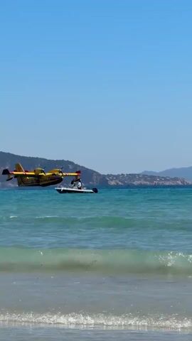 The video captures a dramatic scene on a bright, sunny day at a beach in La Ciotat, France. A large, yellow and red firefighting seaplane, resembling a Canadair CL-415, is seen flying exceptionally low over the clear turquoise waters of the Mediterranean Sea. The aircraft is distinctly marked with "34 SECURITE CIVILE" in black lettering on its side, indicating its affiliation with the French Civil Security.

As the plane skims just meters above the water's surface, it passes directly over a small white boat, which appears to be a paddle boat or dinghy, with one person aboard. Further out in the water, another person's head is visible, swimming or floating. In the foreground, on the sandy beach, a child with long, dark hair stands facing away from the camera, intently watching the seaplane's powerful flight.

The sky is a brilliant, cloudless blue, and small waves gently break on the shore. Distant hills or mountains can be seen across the water in the background. The activity suggests the seaplane is either performing a water-scooping maneuver or executing a very low-altitude flyby, possibly during a training exercise or demonstration. The sounds of excited onlookers can be heard in the background, adding to the palpable sense of awe and excitement generated by the close proximity and impressive display of the aircraft.