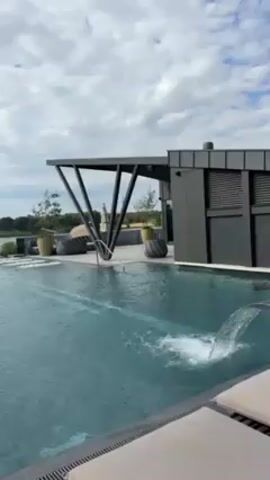 The video shows a swimming pool with a dark-colored tile finish and a small waterfall feature. The pool appears to be an infinity pool, merging with the horizon, which features a body of water. Behind the pool is a modern structure with dark grey siding and a geometric pergola.  Lounge chairs are visible on the pool deck. The sky is partly cloudy, with the clouds reflecting in the pool's surface.