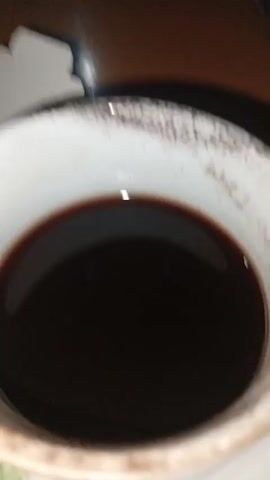 The media file features a close-up shot of a white teacup filled with dark liquid, likely coffee or tea. The teacup has a decorative floral pattern along its base and appears to be slightly worn. It sits on a saucer, also with a floral design. To the left of the teacup, a small, folded white object, possibly a piece of packaging or a lid, is visible. The background suggests an indoor setting, with a polished wooden surface, likely a table, visible behind the teacup. The lighting appears to be artificial, casting shadows and creating a warm glow on the wooden surface. There is no visible text within the media file. The scene is static, with no discernible activity taking place beyond the placement of the teacup and the small object. The overall mood is calm and still.