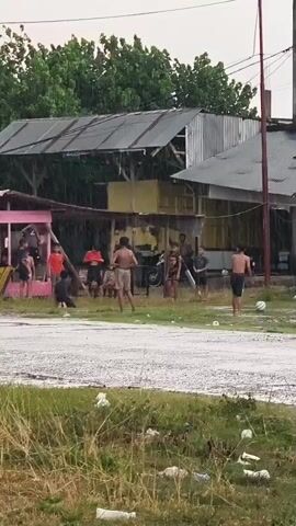 The video shows a group of people playing football in the rain. The scene is outdoors with a road, grass, and several buildings in the background. Some of the buildings appear to be informal structures with corrugated metal roofs. A motorcycle passes by in the foreground.