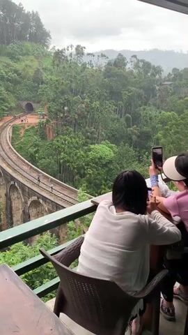 The media captures a scenic view of the Nine Arch Bridge in Ella, Sri Lanka, from a vantage point above and to the side. Three individuals are visible in the foreground, seated at a table on a balcony or terrace, all engrossed in using their mobile phones to take pictures or videos. The person in the middle wears a large visor, shielding their face. The bridge itself is a prominent architectural feature, an arched railway viaduct constructed with stone and bricks, curving through a lush, verdant landscape.

The surrounding environment is characterized by dense tropical foliage, with numerous tall palm trees and other greenery blanketing the hills. A railway line runs across the bridge, and several people are seen walking along the tracks, some holding umbrellas, suggesting a light rain or recent shower. A tunnel entrance is visible on the hillside where the railway emerges from the dense vegetation. The sky appears overcast, indicating a cloudy or potentially rainy day. The overall atmosphere suggests a tourist attraction, with visitors documenting the iconic landmark. The time of day is likely daytime, given the ambient light. No visible text is present in the media.