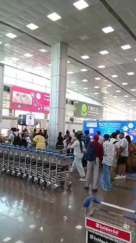 The video shows a busy airport terminal. Many people are standing around, some with luggage or pushing luggage carts. The floor is shiny and reflective. There are multiple advertising signs on the walls. The ceiling is high and covered with square fluorescent lights.  Large white pillars are supporting the ceiling. The number 4 is prominently displayed in multiple locations.