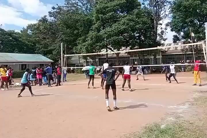 The video shows a volleyball game in progress, taking place on a dirt court outdoors. There are approximately 12 players on the court, divided into two teams. The players are wearing athletic attire of different colors including white, red, green, and black.  A white net separates the two sides of the court. Spectators are standing on the sidelines, watching the game. There are buildings and trees in the background. The weather appears sunny with some cloud cover. The volleyball is in the air. Players are actively moving and attempting to hit the ball.