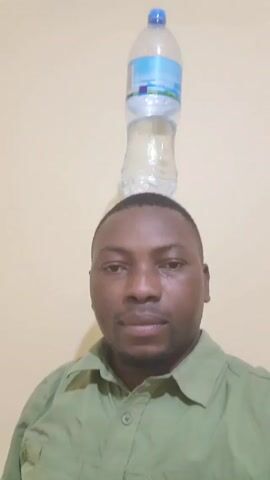 A man with short hair and a green collared shirt looks directly at the camera. He has a water bottle balanced on his head. The background is a plain, light-colored wall.