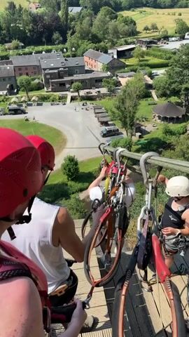 This is a vertical, point-of-view video filmed from a high vantage point, looking down on a ziplining course. In the foreground, three individuals wearing red and white helmets and harnesses are preparing to zipline. Their backs are to the camera. To the right, a fourth person, wearing a white helmet, is positioned on the platform, holding onto the zipline equipment.

The zipline cable stretches out over a lush green valley, dotted with trees and a winding river. Below the zipline, a small village with traditional brick buildings and a parking lot is visible. There are several cars parked in the lot. A large, dark, tent-like structure is also present in the village area. The scene appears to be a recreational activity in a rural setting.

The time of day is clearly daytime, with bright sunlight casting shadows. The sky is a clear, vibrant blue, indicating good weather. The overall mood seems to be one of excitement and anticipation, as the individuals are about to embark on an adventurous activity. The video captures the natural beauty of the Belgian countryside. There is no visible text in the video.